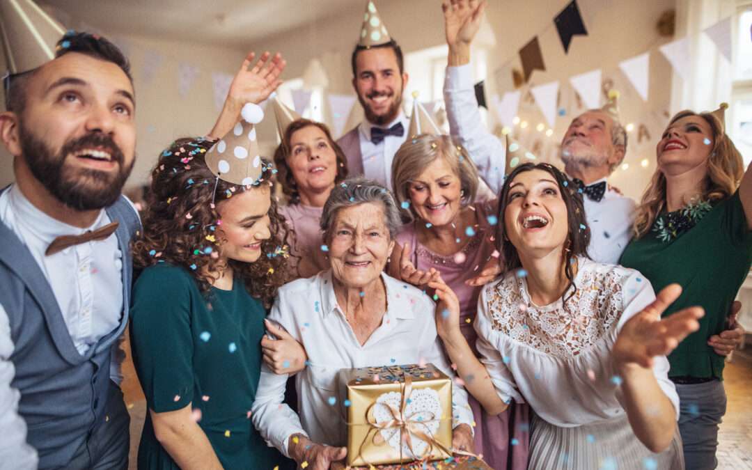 A happy senior woman surrounded by family and friends celebrating her birthday with party hats, confetti, and a gift at a festive birthday party for seniors.