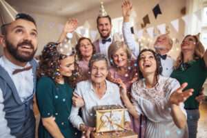 A happy senior woman surrounded by family and friends celebrating her birthday with party hats, confetti, and a gift at a festive birthday party for seniors.