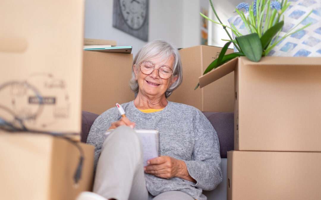 Senior woman sitting among moving boxes, smiling and writing in a notebook as she plans her move.