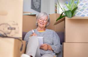 Senior woman sitting among moving boxes, smiling and writing in a notebook as she plans her move.